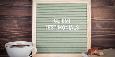 Felt letter board displaying “CLIENT TESTIMONIALS,” set on a wooden surface beside a coffee cup, books and a stamp.